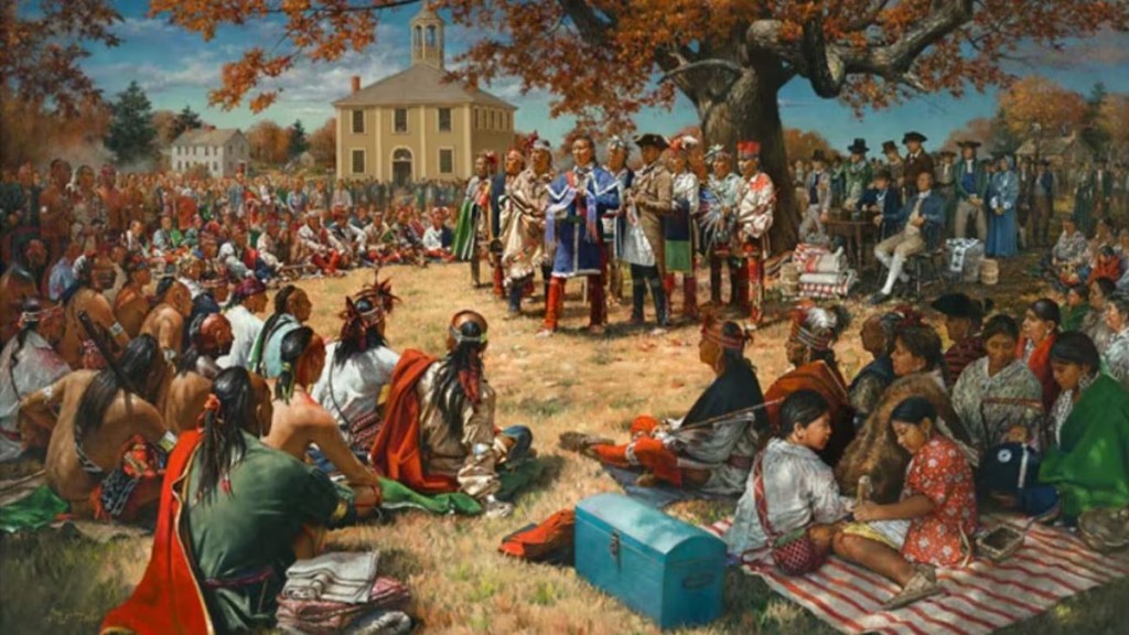 Un rassemblement historique avec des membres de différentes communautés indigènes, en tenue traditionnelle, écoutant un discours près d'un grand arbre, avec un bâtiment en arrière-plan.